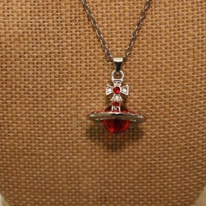 Peut inclure: Collier argenté avec un pendentif représentant un globe rouge et une croix. Le pendentif est entouré d'un anneau argenté. Le collier est présenté sur un fond texturé de couleur beige.