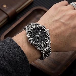 May include: A silver-toned wristwatch with an ornate, textured bezel and band. The watch face is black with a cross design. The watch is on a wrist, with a leather wallet and wooden tray in the background.