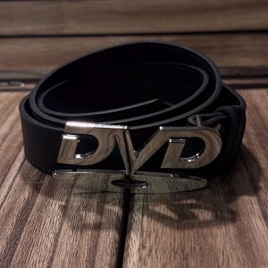 Könnte beinhalten: Schwarzer Ledergürtel mit silberner Schnalle in Form der Buchstaben "DVD". Der Gürtel ist auf einer Holzoberfläche aufgerollt.