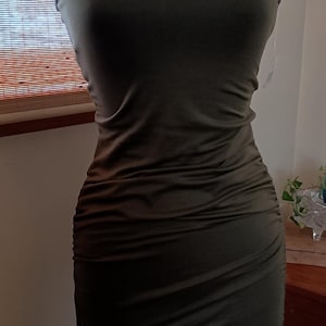May include: Olive green bodycon dress with spaghetti straps. The dress is ruched on the sides and is displayed on a black mannequin. The dress is a solid color and appears to be made of a soft, stretchy material. The dress is form-fitting.
