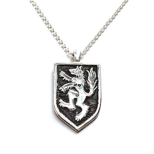 Op de afbeelding: Een zilveren ketting met een hanger in de vorm van een schild. Het schild heeft een zwarte achtergrond en een zilveren wolfontwerp.