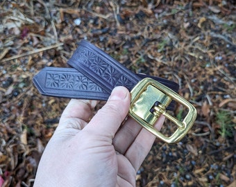 May include: A brown leather belt with a gold metal buckle. The belt has a floral pattern embossed into the leather.