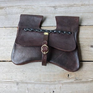 Pode incluir: Uma bolsa de couro marrom com uma aba e um fecho de fivela. A bolsa tem uma alça trançada e um pequeno bolso na frente.