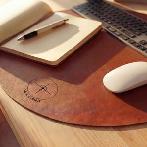 Könnte beinhalten: Eine braune Leder-Schreibtischunterlage mit abgerundeter Kante, mit einem kreisförmigen Logo und dem Namen "WILLIAM". Ein Notizbuch, Stift, Tastatur und weiße Maus liegen auf der Unterlage, auf einem Holztisch.