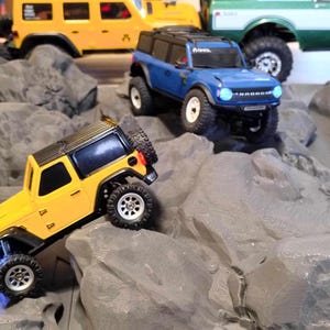 May include: Miniature toy off-road vehicles in yellow and blue, traversing a rocky landscape. A yellow Jeep-style vehicle is in the foreground, with a blue Ford Bronco and other vehicles in the background.