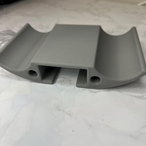 Op de afbeelding: Een grijs, 3D-geprint object met een rechthoekig middengedeelte en gebogen, halfronde uiteinden. Twee gaten zijn zichtbaar aan de onderkant. Het object lijkt een houder of ondersteunende structuur te zijn, mogelijk voor elektronica of andere kleine items.