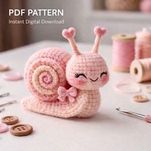 Pode incluir: Um caracol rosa de crochê com uma concha rosa claro e bege, antenas em forma de coração e um pequeno laço. O caracol tem um rosto sorridente com bochechas rosadas. O texto na parte superior diz "PDF PATTERN Download Digital Instantâneo".