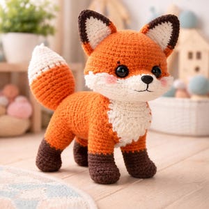 May include: A crocheted fox toy in orange, white, and brown. The fox has a white chest and tail tip, with brown accents on the ears and feet. It has black eyes and pink cheeks. The toy is standing on a wooden surface.