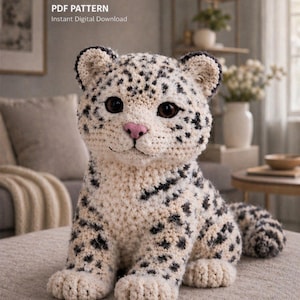 May include: A crocheted snow leopard plush toy with black spots and a pink nose. The text "PDF PATTERN Instant Digital Download" is visible in the upper left corner. The handmade animal is sitting on a neutral-colored surface.