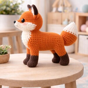 May include: A crocheted fox figurine in orange, white, and brown. The fox has a white chest, a white-tipped tail, and brown accents on its ears and feet. It stands on a light wooden table. The fox is a handmade item.