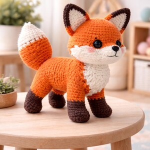 May include: A crocheted fox figurine in orange, white, and brown. The fox has large black eyes, a white chest, and a fluffy white-tipped tail. It stands on a small wooden table. The fox is a handmade item.