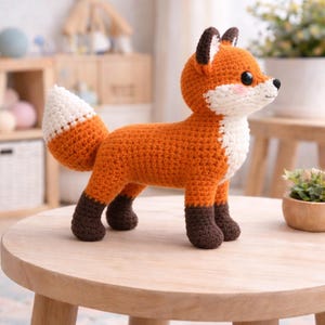 May include: A handmade crochet fox toy in orange, white, and brown. The fox has a fluffy white tail, dark brown paws, and a cute face with black eyes and pink cheeks. It is sitting on a small wooden table.