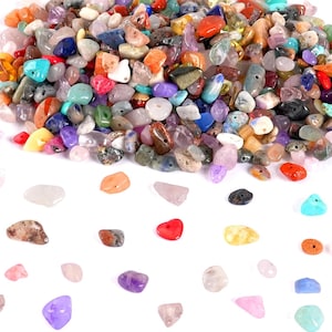 May include: A collection of colorful, polished gemstones in various shapes and sizes. The stones are a mix of colors, including red, orange, blue, green, purple, and clear. Some stones have holes drilled through them. The gemstones are scattered on a white surface.
