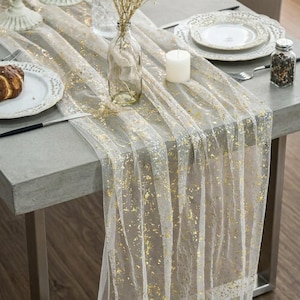 May include: A sheer white table runner featuring scattered gold foil flakes. The runner is draped across a gray table, accompanied by a clear glass vase with dried flowers, a white pillar candle, white plates, and silver cutlery.