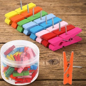 May include: A collection of multi-colored wooden clothespins with attached metal push pins. The set includes a clear plastic storage jar filled with assorted colorful clips and a single orange clip displayed upright on its side.