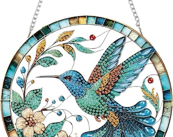 Puede incluir: Un atrapasoles circular que presenta un colibrí en vuelo rodeado de flores y follaje. El diseño está compuesto por pequeñas cuentas facetadas multicolores en tonos azul, verde, dorado y crema, dentro de un marco de metal oscuro.