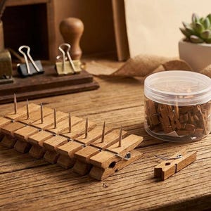 May include: A row of wooden clothespins with metal pins inserted into the top, a clear plastic jar filled with additional wooden clothespins, and a single loose wooden clothespin resting on a rustic wooden surface with office supplies.