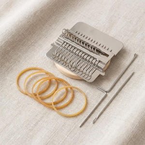 May include: A metal darning loom tool featuring a rectangular plate with multiple hooks, a wooden base, four tan rubber bands, and a long metal needle. This set is designed for textile repair and includes all visible components for mending fabric.
