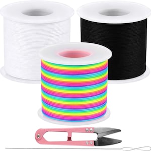 May include: Three spools of elastic cord in white, black, and a multi-colored rainbow stripe pattern. The spools are arranged with the rainbow spool in front. A pair of pink metal thread snips and a thin metal beading needle are included below.