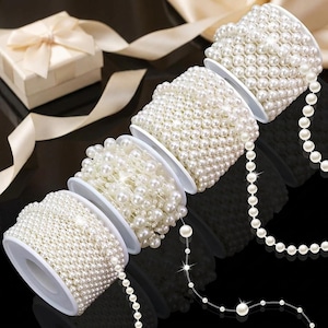 May include: Four white plastic spools wrapped with strands of small, round, cream-colored faux pearls. The pearls are arranged in dense, uniform rows on each spool, with loose strands of pearls extending outward from the spools onto a black surface.