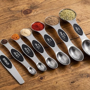 May include: A set of six stainless steel measuring spoons and a leveler tool. Each spoon features a black silicone grip and is labeled with its capacity, ranging from 1/8 tsp to 1 tbsp. The spoons contain various spices and herbs.