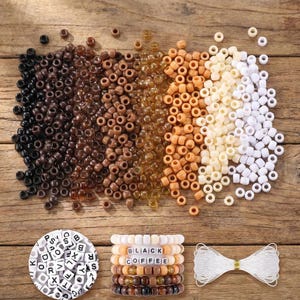 May include: A collection of small, round, pony beads arranged in a color gradient from black to brown, amber, tan, cream, and white. Below are a circular container of white alphabet letter beads, a stack of beaded bracelets, and white string.