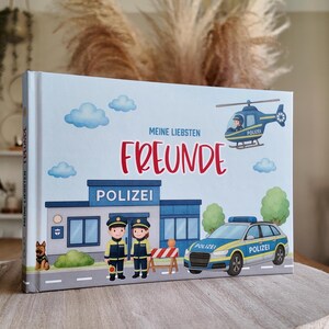 Könnte beinhalten: Ein Kinderbuch mit hellblauem Einband mit Illustrationen von Polizisten, einem Polizeiauto, einem Hubschrauber und einer Polizeistation. Der Titel "Meine Liebsten Freunde" ist rot geschrieben. Das Buch liegt auf einer hellen Oberfläche.