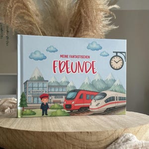 Könnte beinhalten: Ein Kinderbuch mit hellblauem Einband, das eine Bahnhofsszene zeigt. Der Titel "MEINE FANTASTISCHEN FREUNDE" ist rot. Die Illustration zeigt Züge, Wolken, Berge und eine Uhr.