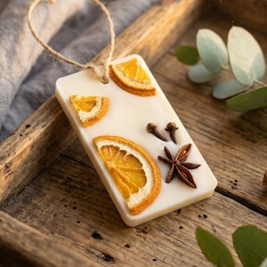 May include: A rectangular, off-white wax melt with dried orange slices, cloves, and star anise. A natural twine loop is attached for hanging. The wax melt is displayed on a wooden tray, suggesting a home fragrance product.