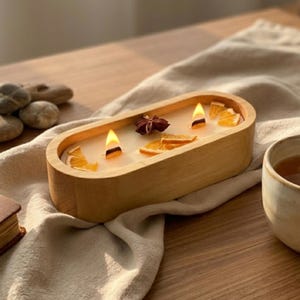 May include: An oval-shaped wooden candle vessel containing white wax with two lit wicks. The surface of the wax is decorated with dried orange slices and a single star anise. The candle is positioned on a light-colored fabric cloth.