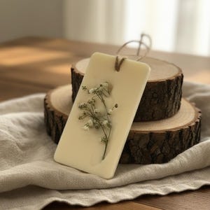 May include: A rectangular, off-white air freshener with a sprig of delicate white flowers embedded within. It hangs from a natural twine loop. The freshener rests on a wooden slice, creating a rustic, natural aesthetic.
