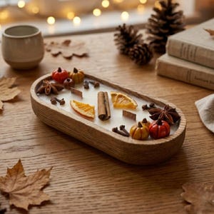 Puede incluir: Una vela ovalada en un cuenco de madera rellena de cera blanca, decorada con rodajas de naranja seca, ramas de canela, anís estrellado, clavos de olor y pequeñas calabazas decorativas naranjas y rojas con dos mechas de madera.