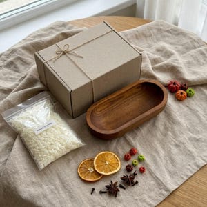 May include: A brown cardboard gift box tied with twine, a clear plastic bag containing white natural soy wax flakes, an oval wooden bowl, two dried orange slices, star anise, and small decorative pumpkins on a beige linen cloth.