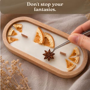 May include: An oval wooden candle container filled with white wax, featuring two wooden wicks, dried orange slices, and a star anise being placed with metal tweezers. The text "Don't stop your fantasies." is visible at the top of the image.