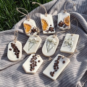May include: A selection of handmade white wax melts in various shapes. Some are decorated with dried orange slices, star anise, and cloves. Others feature coffee beans or small white flowers. Each melt is strung with jute twine.