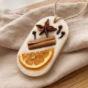 Può includere: Un ornamento in cera bianca di forma ovale con elementi botanici incorporati, tra cui una fetta di arancia essiccata, due bastoncini di cannella, un anice stellato e tre chiodi di garofano. Un sottile laccio in juta è fissato in alto.