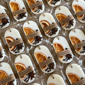 Puede incluir: Adornos ovalados de cera blanca decorados con una rodaja de naranja seca, un anís estrellado y clavos de olor. Cada adorno está envuelto en plástico transparente con una banda de papel marrón impresa con el texto Thank you for joining Shaya's Bar Mitzva!