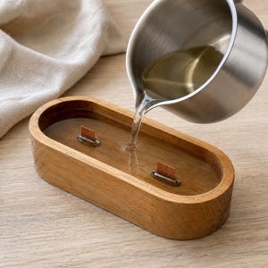 May include: A stainless steel pitcher pours clear liquid wax into an oval-shaped wooden candle vessel. The vessel features two rectangular metal wick tabs secured at the base, waiting to be filled with the molten wax for candle making.