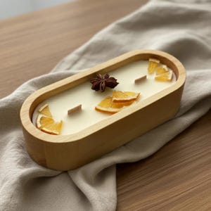 May include: An oval-shaped wooden candle bowl filled with cream-colored wax. The candle features two flat wooden wicks, several dried orange slices, and a single star anise pod embedded on the surface as decorative elements.