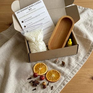 May include: A DIY wooden bowl candle kit in a cardboard box containing a wooden oval bowl, a bag of white wax pellets, a yellow dropper bottle, two metal wick tabs, and an instruction sheet. Dried orange slices, star anise, and cloves are included.
