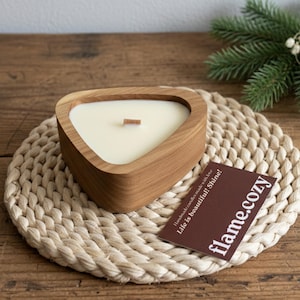 Pode incluir: Um castiçal triangular de madeira com uma vela branca cremosa e um pavio de madeira. A vela está sobre um descanso de mesa tecido. Um cartão castanho com o texto "flame.cozy" é visível.
