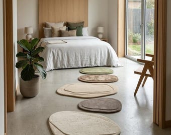 May include: A series of five irregular, pebble-shaped area rugs arranged in a line on a concrete floor. The rugs feature varying neutral tones including cream, beige, brown, and sage green, each with a raised, layered texture and soft edges.