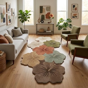 May include: A floor rug shaped like a cluster of overlapping hibiscus flowers in shades of cream, olive green, terracotta, tan, sage green, and dark brown, featuring textured petals and central stamen details, laid out on a light wood floor.