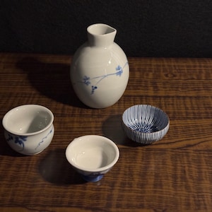 以下が含まれることがあります： 青い花の模様が入った白い陶器の酒器セット。セットには丸みを帯びたボトルと、青いストライプのカップを含む3つの小さなカップが含まれています。木製の表面に配置されています。