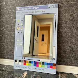 Può includere: Uno specchio rettangolare incorniciato per assomigliare all'interfaccia dell'applicazione Windows Paint. La cornice azzurra presenta una barra degli strumenti con icone e il testo "mel's world - Paint". Lo specchio riflette una stanza con porte aperte e un muro grigio scuro.