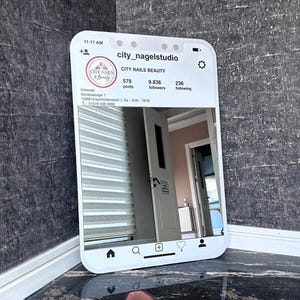 Puede incluir: Un espejo rectangular diseñado para parecerse a una página de perfil de redes sociales de City Nails Beauty. El marco presenta bordes blancos con iconos, recuento de seguidores y dirección. La superficie refleja una habitación con una puerta.