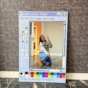 Può includere: Uno specchio rettangolare incorniciato per assomigliare alla classica interfaccia del programma Paint di Windows. La cornice azzurra presenta una barra degli strumenti con icone, una tavolozza di colori e il testo "mel's world - Paint". Lo specchio riflette una persona che scatta una foto.