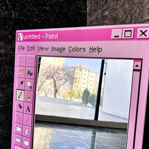 Puede incluir: Un espejo enmarcado en rosa dise&ntilde;ado para parecerse a la ventana de la aplicaci&oacute;n "Paint" cl&aacute;sica. El espejo presenta una barra de men&uacute; con "File, Edit, View, Image, Colors, Help" y una barra de herramientas con varias herramientas de dibujo. El espejo refleja una vista de un edificio y &aacute;rboles.