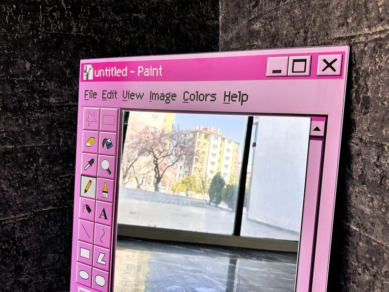 Puede incluir: Un espejo enmarcado en rosa dise&ntilde;ado para parecerse a la ventana de la aplicaci&oacute;n "Paint" cl&aacute;sica. El espejo presenta una barra de men&uacute; con "File, Edit, View, Image, Colors, Help" y una barra de herramientas con varias herramientas de dibujo. El espejo refleja una vista de un edificio y &aacute;rboles.