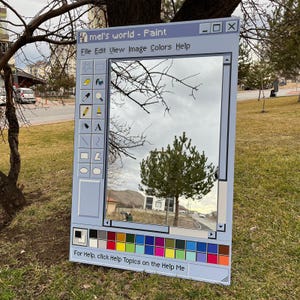 Könnte beinhalten: Ein großer, rechteckiger Spiegel, der wie eine klassische Computer-Paint-Programmoberfläche gestaltet ist. Der Rahmen des Spiegels ist hellblau, mit einer Symbolleiste und einer Farbpalette an den Seiten. Der Spiegel reflektiert einen bewölkten Himmel, Bäume und ein Gebäude.